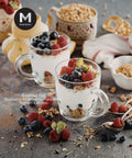 Two clear glass mugs filled with layered breakfast yogurt cups topped with blueberries, strawberries, raspberries, and granola, arranged on a rustic dark surface with additional berries, muesli, and a teapot in the background. White text on the left reads 'Breakfast Cup Blueberry, Strawberry, Raspberry Yoghurt & Muesli' and a black circular MMMMI logo appears near the top left.