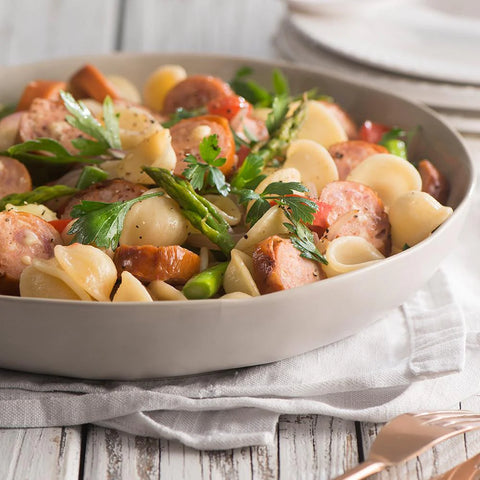 Pasta dish with Big Cheese Kransky sausages and vegetables in a white bowl on a wooden table.