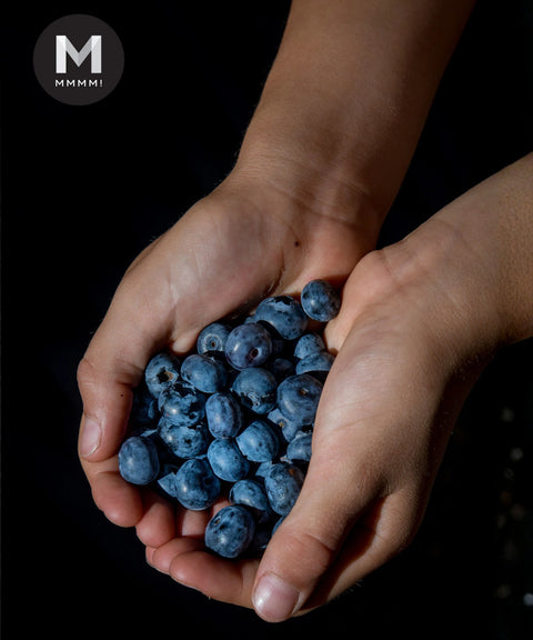 Two hands cupping a small pile of fresh blueberries against a black background, with a circular 'MMMMI' logo in the top-left corner.