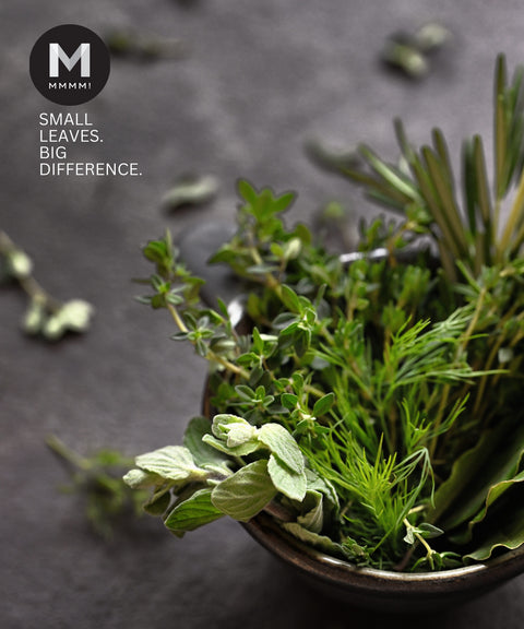 Bowl of green herbs on a dark surface with 'Mmmm' brand logo.