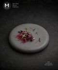 A round gray stone plate on a dark textured background, topped with a small pile of red amaranth microgreens. The MMMMI logo appears in the top-left corner, and the text 'Red Amaranth Microgreens' appears in the bottom-right. Tiny red leaves is perfect for garnish.