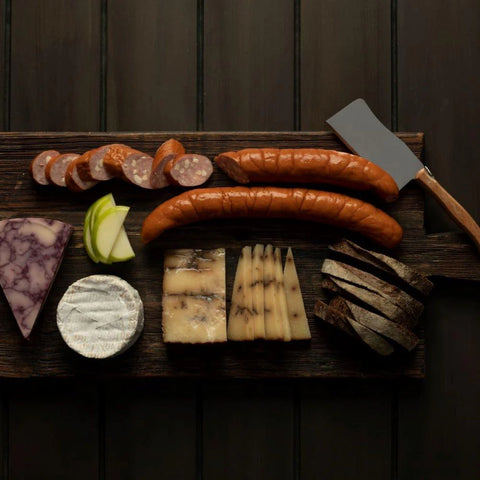  Götzinger Cheese kabana with assorted cheeses, and bread on a wooden board with a knife against a dark background.
